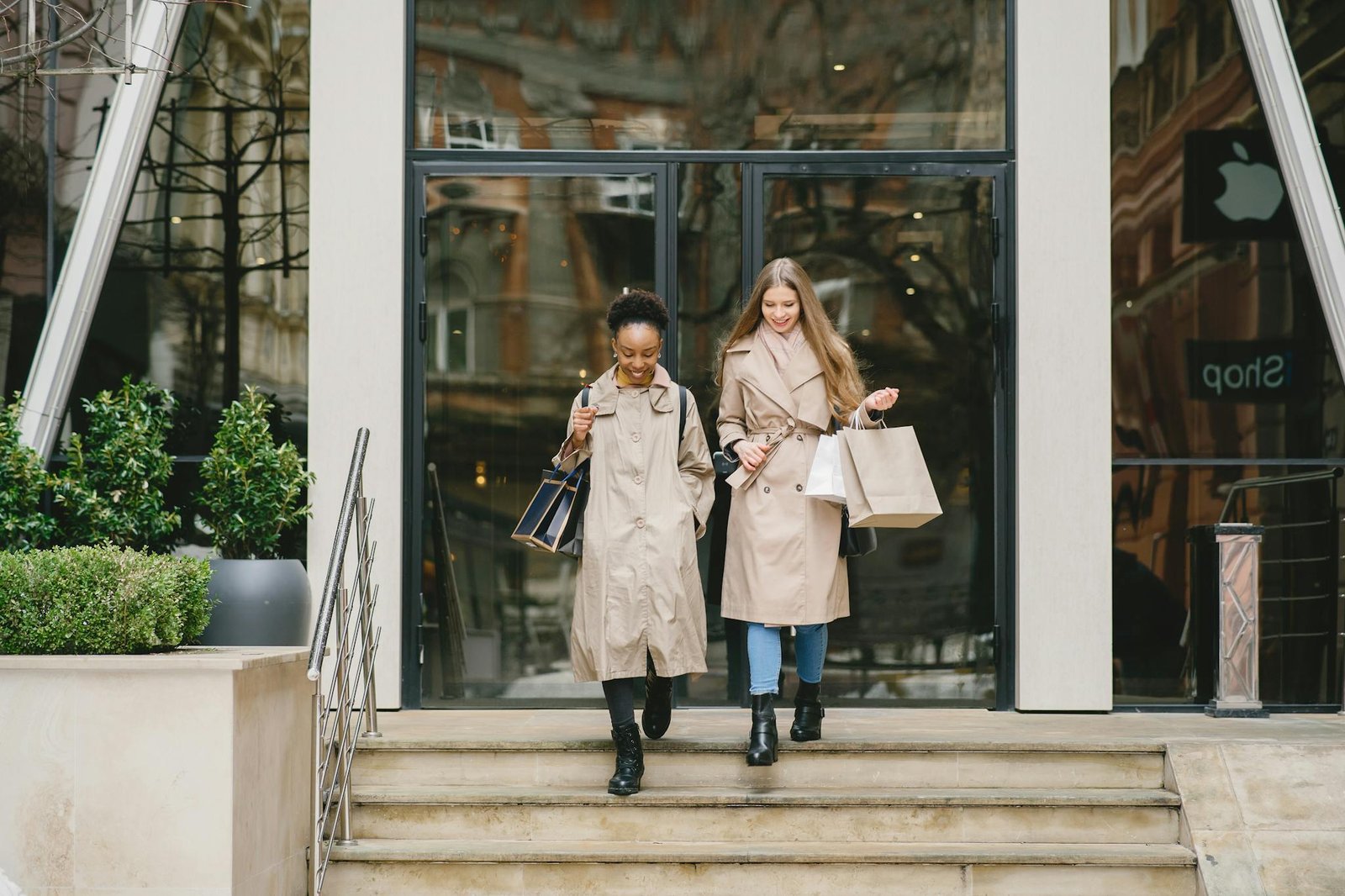 Two women in trench coats carrying shopping bags exit a store, showcasing modern fashion and style.